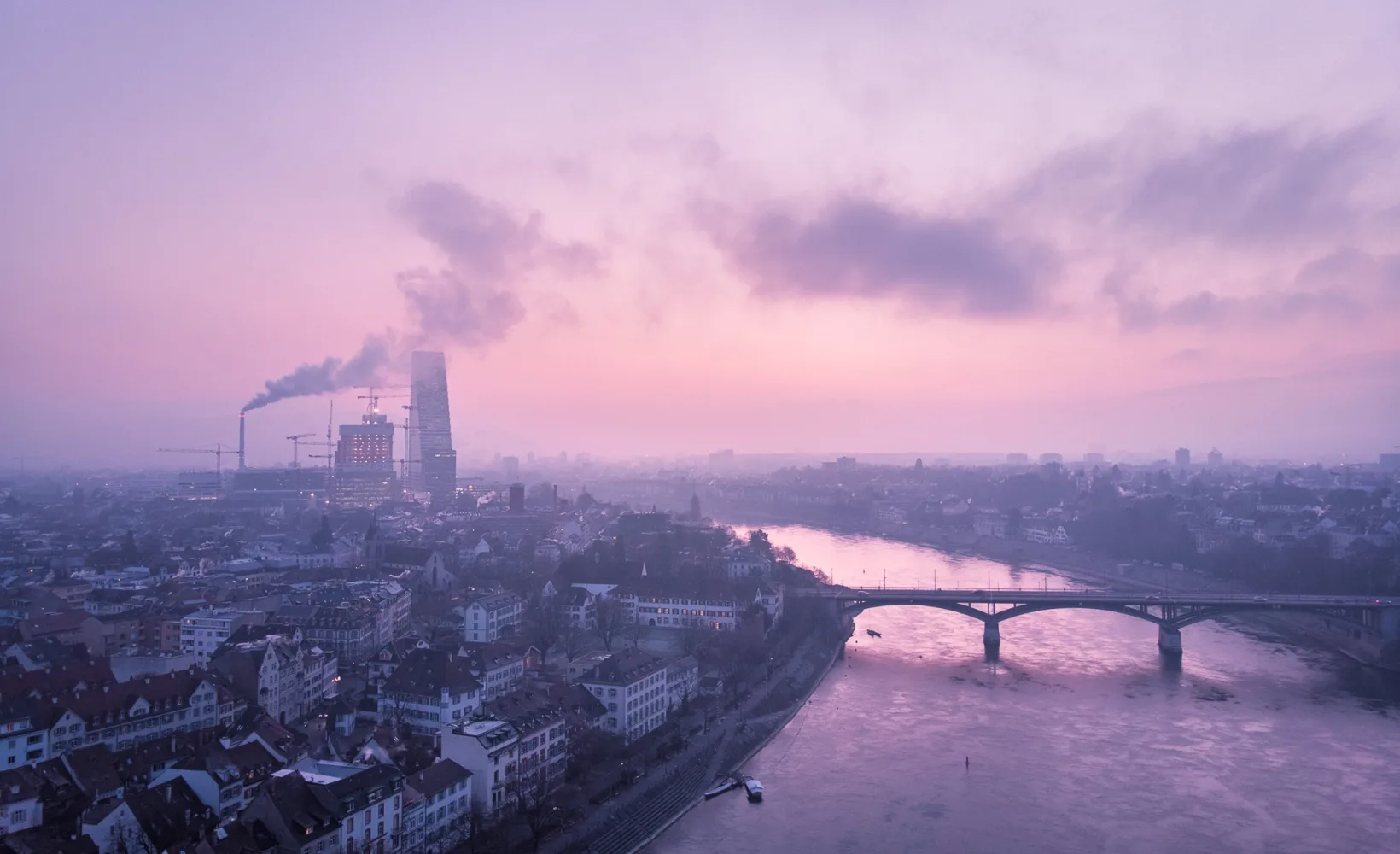 Blick aus der Vogelperspektive auf Basel. Zu sehen sind die Roche-Türmie, die Wettsteinbrücke und der Rhein. Der Himmel und das gesamte Foto ist in pink und blauen Farbtönen.