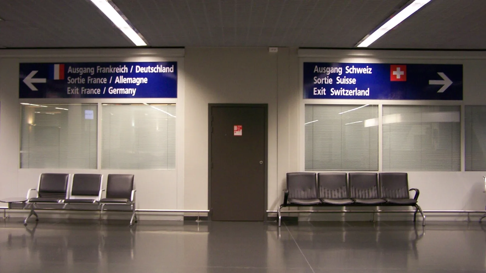 Zwei Schilder für Ausgänge, die je nach links (Frankreich) oder rechts (Schweiz) zeigen. Davor Stühle an einer Wand.