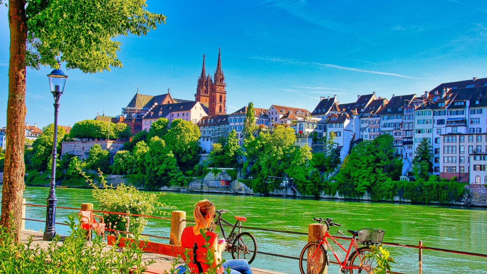 Sehr farbiges Foto des Basler Münsters, davor der Rhein und mehrere am Geländer abgestellte Velos.