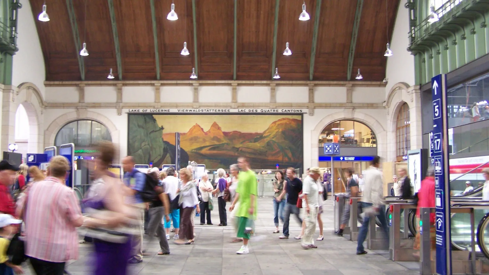 Foto der Bahnhofshalle des Bahnhofs Basel SBB. Viele Menschen (verschwommen) bewegen sich über grauen Steinboden vor einem grossen Gemälde des Vierwaldstättersees.