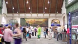 Foto der Bahnhofshalle des Bahnhofs Basel SBB. Viele Menschen (verschwommen) bewegen sich über grauen Steinboden vor einem grossen Gemälde des Vierwaldstättersees.