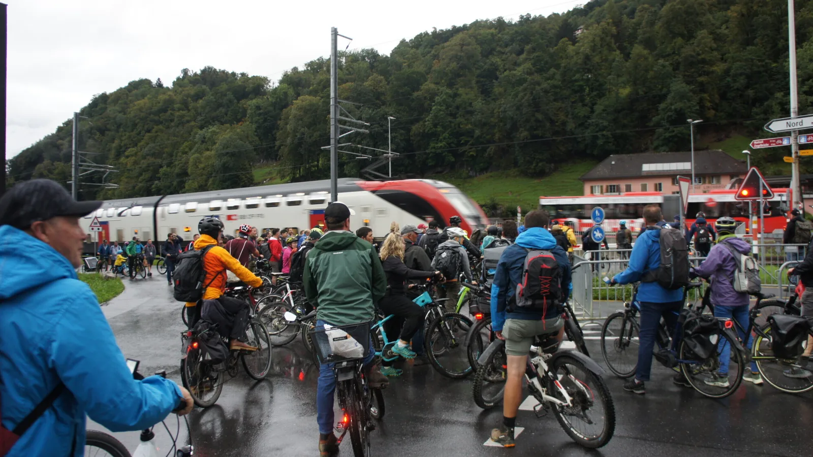 Im Vordergrund stauen sich Velofahrende vor einem geschlossenen Bahnübergang. Im Hintergrund sichtbar sind ein FV-Dosto Zug und ein Bus