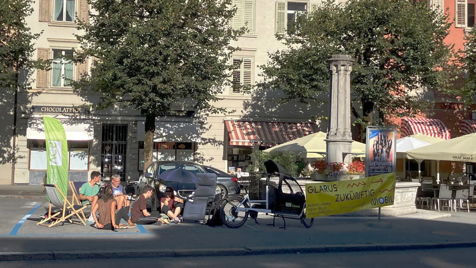 Sicht von der gegenüberliegenden Strassenseite auf die zwei besetzten Parkplätze. Mehrere Leute sitzen auf Stühlen oder dem Boden. Links die VCS Fahne, rechts ein Lastenrad und Banner